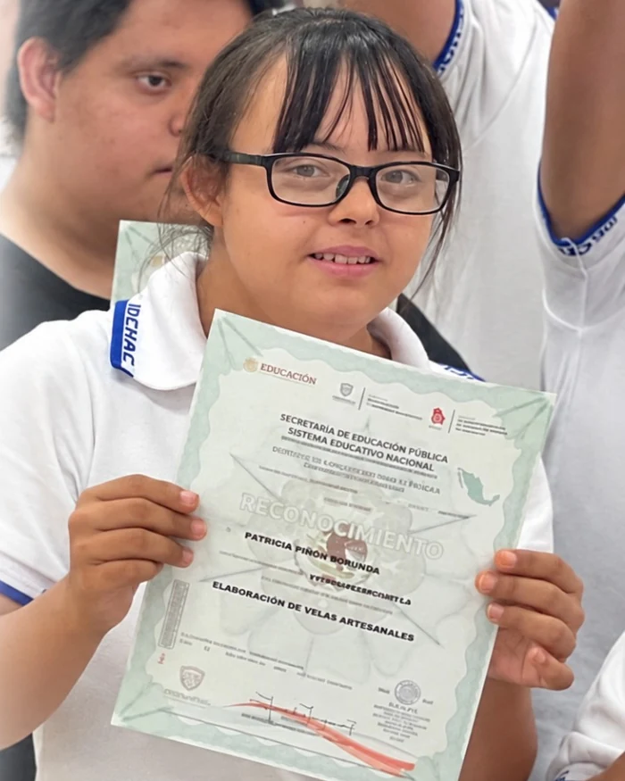 Paty, con lentes y uniforme escolar, sostiene un reconocimiento.